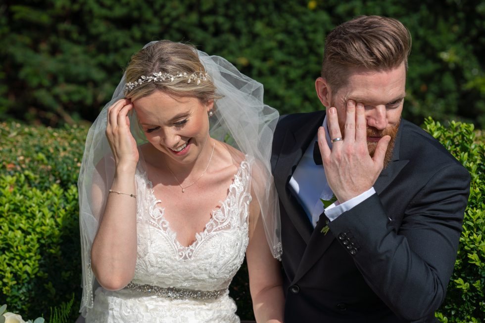 Bride in a tiara and veil with her hand to her forehead beside a groom wiping his eye in front of a hedge
