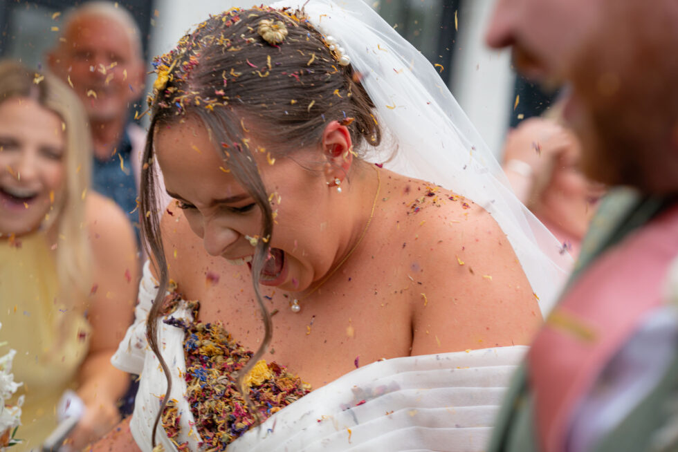 Bride laughing with her head down as a whole box of floral confetti is thrown over her