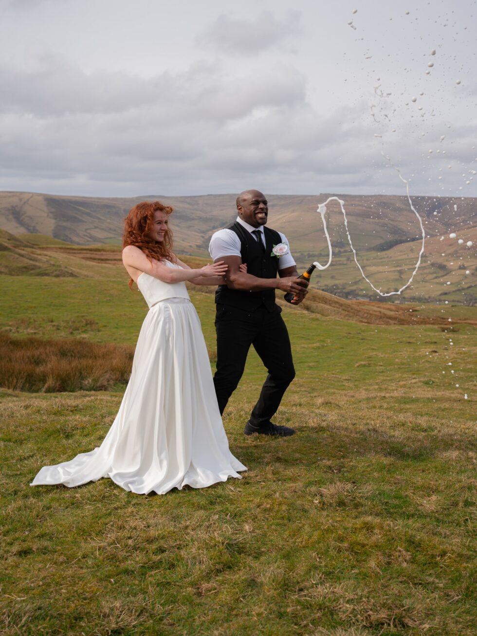 Bride in a white dress and groom in a black waistcoat popping champagne on a grassy hillside under a cloudy sky