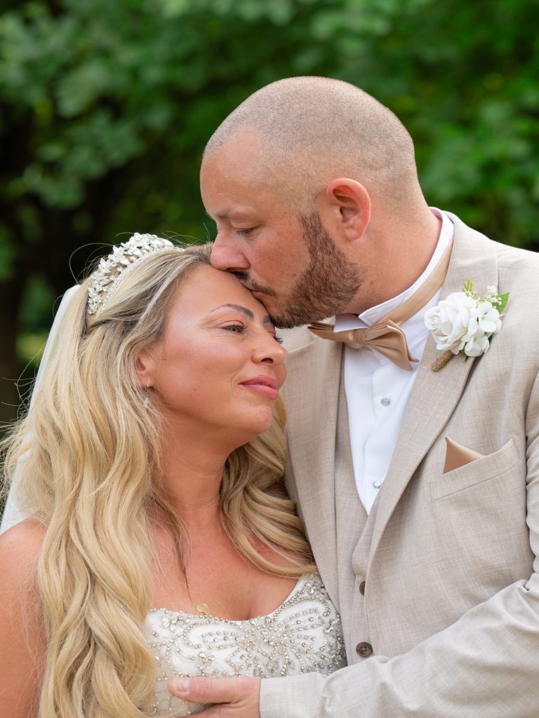 Groom in a stone-coloured suit and bow tie kissing his bride on the forehead, her tiara and long wavy blonde hair framed by green foliage