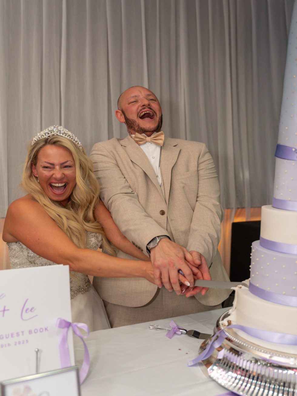 Bride in a tiara and groom in a beige suit laughing hard as they cut their tall wedding cake together