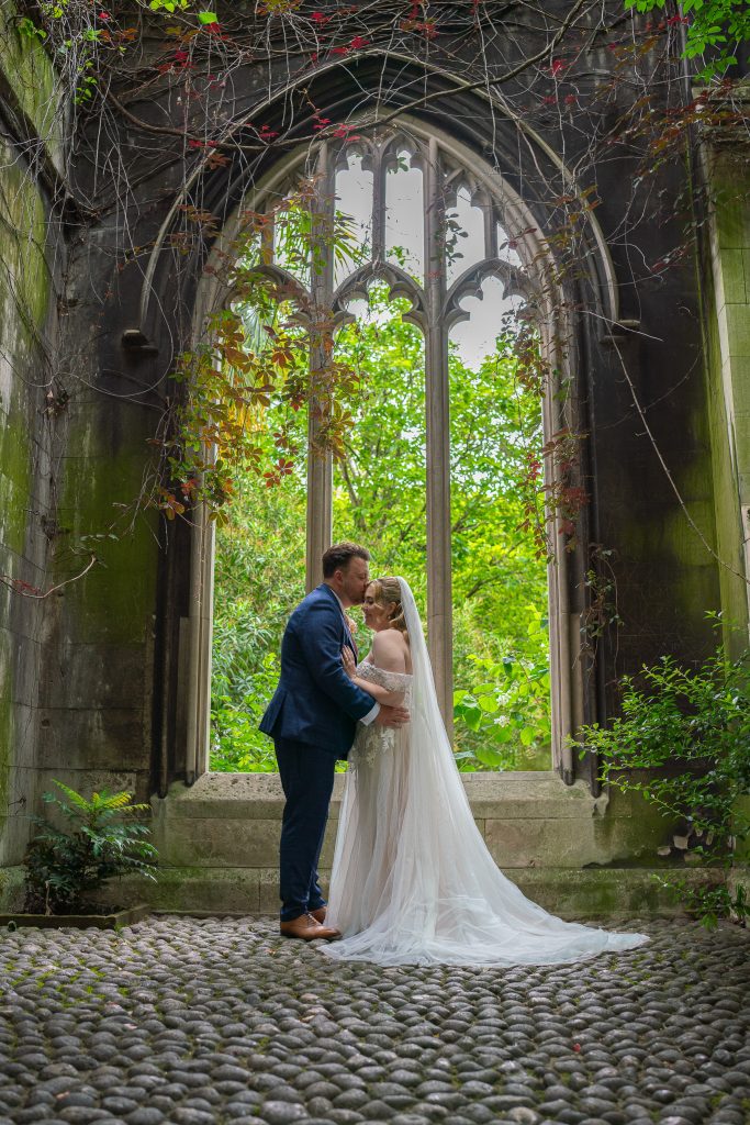 Groom in a blue suit kissing his bride on the forehead inside a ruined stone arch with a tall Gothic window, overgrown with vines, her long veil trailing over a cobbled floor