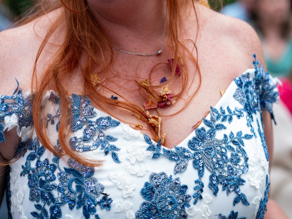 Close-up detail of the bride's white and blue embroidered wedding dress, showing strands of red hair and dried flower confetti stuck in the neckline after the shower at Enchanted Garden Weddings. Candid detail photography.