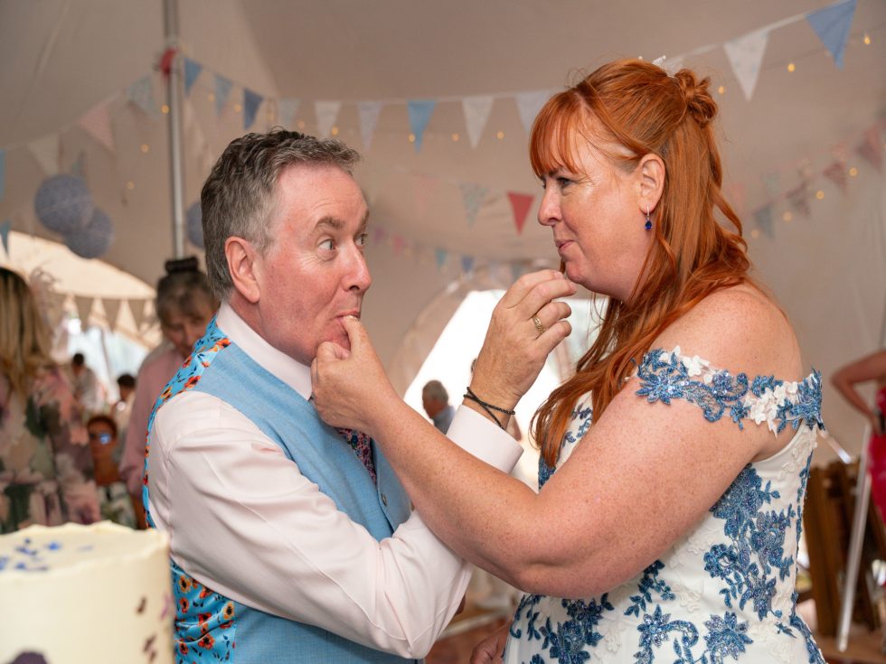 Two wedding guests licking icing off their fingers beside a cake under a tent decorated with bunting