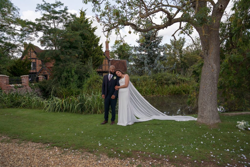 Bride resting her head on her groom’s shoulder beside a pond, her long cape train trailing across the grass, with a timber-framed building and a large tree behind them