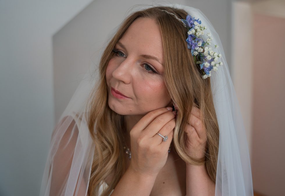 Close-up portrait of the bride putting on her wedding earrings as the final touch during bridal prep, wearing a floral crown and veil. Captured with soft, natural light by a Bedfordshire wedding photographer.