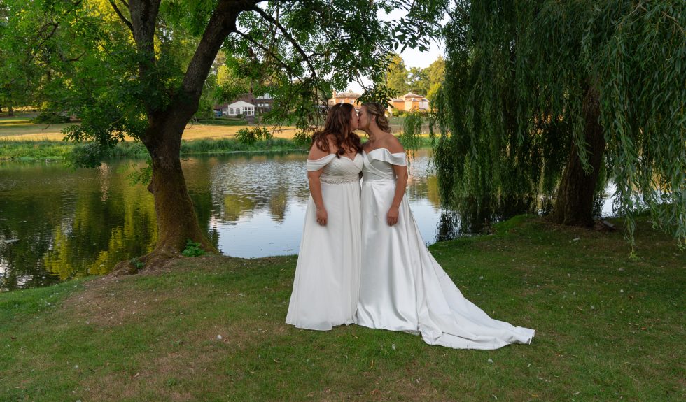 A romantic wedding portrait of two brides sharing a tender kiss under weeping willow trees by a calm lake. Captured by a diverse and inclusive Hertfordshire wedding photographer celebrating the day LGBTQ+ females marry.