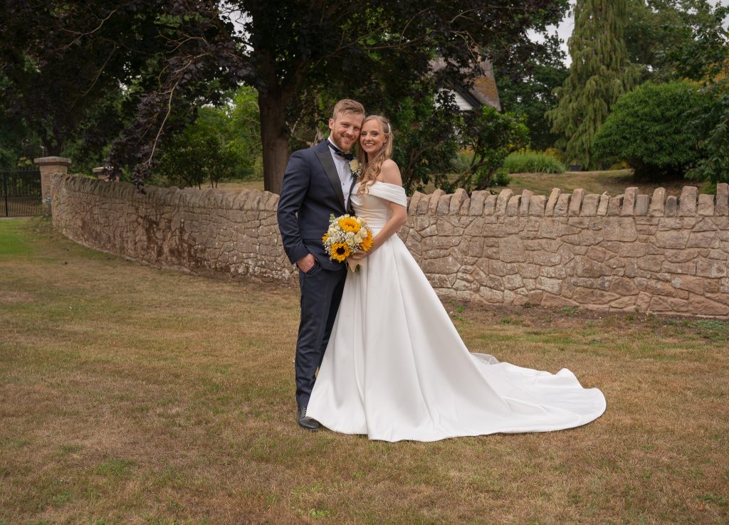 Smiling groom in a navy dinner suit and bow tie and bride in an off-the-shoulder white gown holding a sunflower bouquet, standing cheek to cheek in front of a dry stone wall and trees