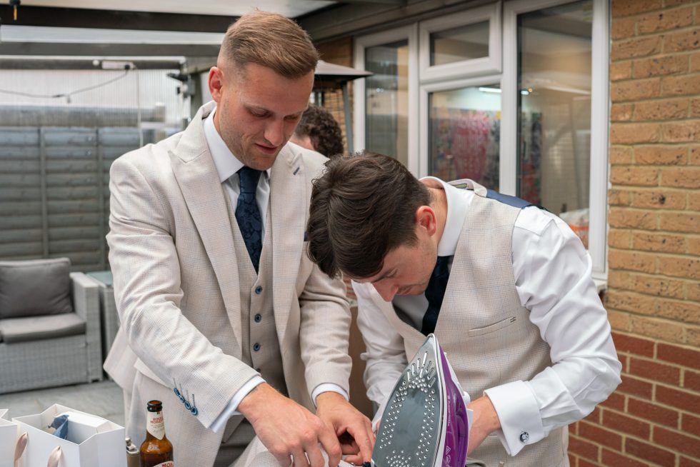 Candid photo of groomsmen helping each other iron a shirt and finalize suit details during the morning prep. Captured by a documentary Hertfordshire wedding photographer.