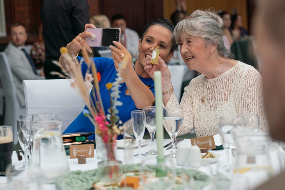 Two wedding guests, an older woman and a younger woman, smiling and taking a cheerful selfie at a beautifully set reception table during the wedding breakfast.