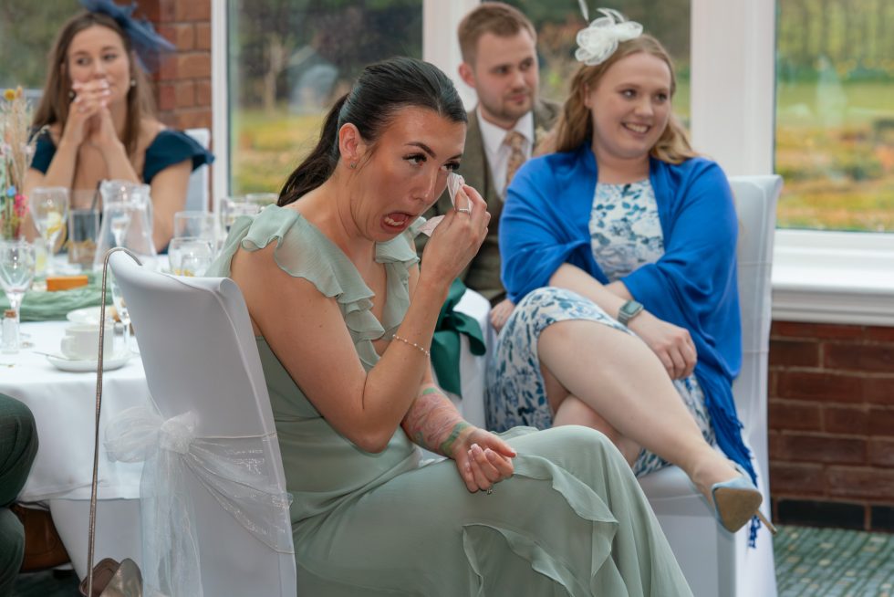 Candid photo of a bridesmaid wiping away a tear while crying emotionally during the wedding reception speeches, seated at a beautifully set table.