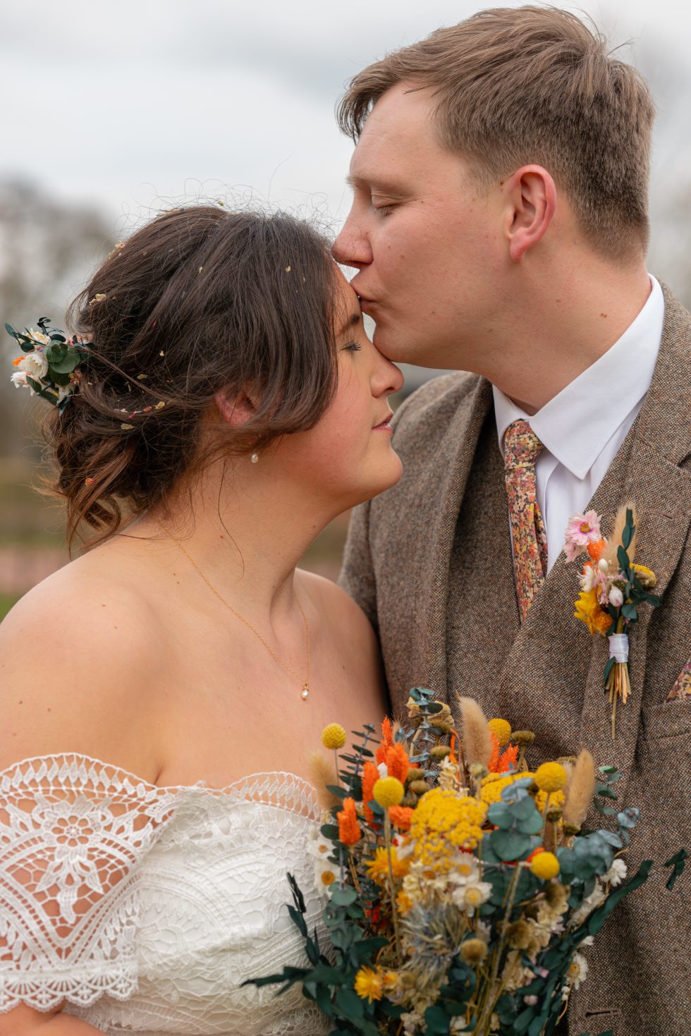 Intimate portrait of a groom kissing his bride on the forehead, featuring a tweed suit and a bright dried flower bouquet by a Hertfordshire wedding photographer.