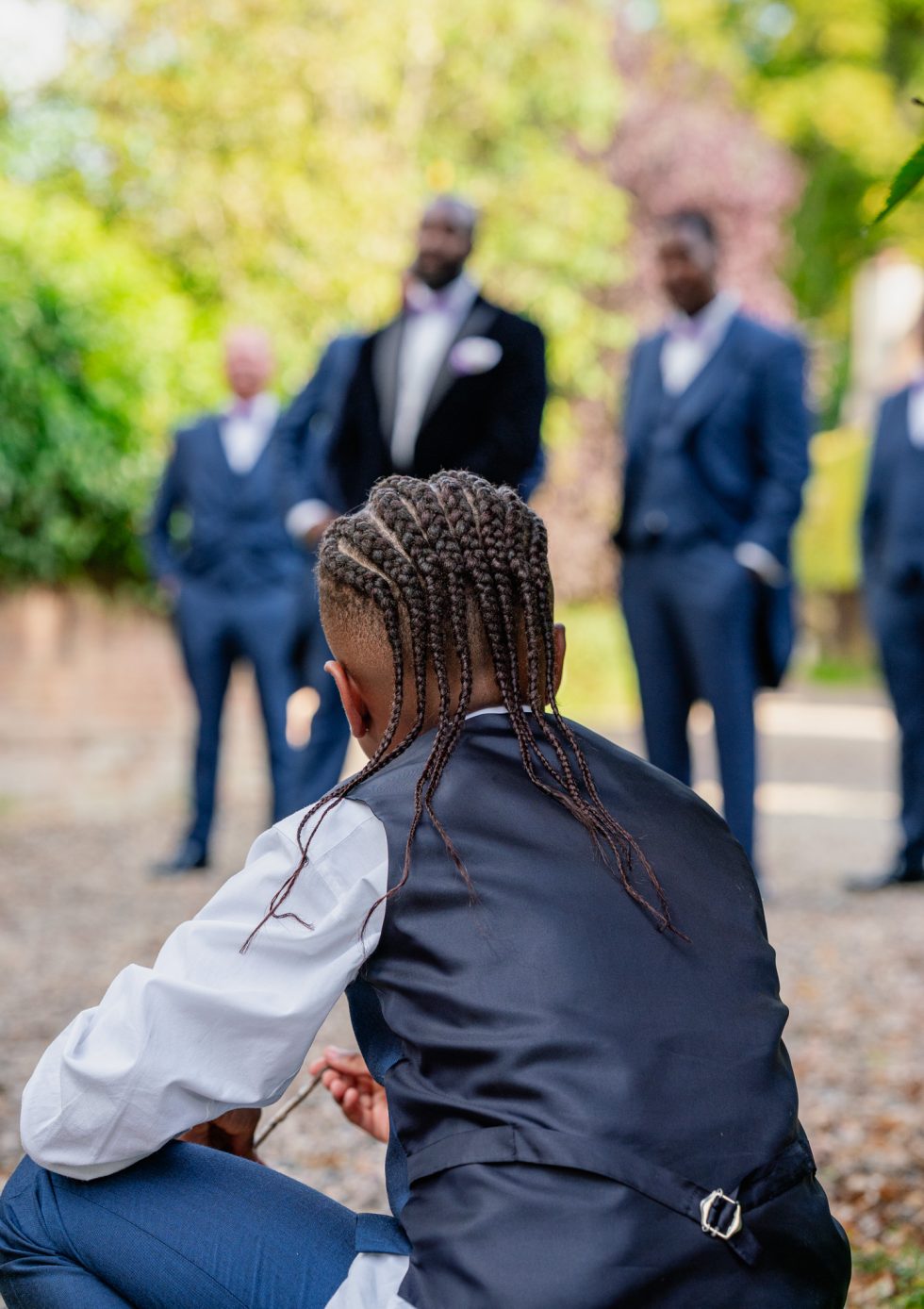 A young wedding guest with braided hair, wearing a navy waistcoat and white shirt, sits in the foreground. In the blurred background, a groom and his groomsmen in navy suits stand in a sunlit outdoor garden.