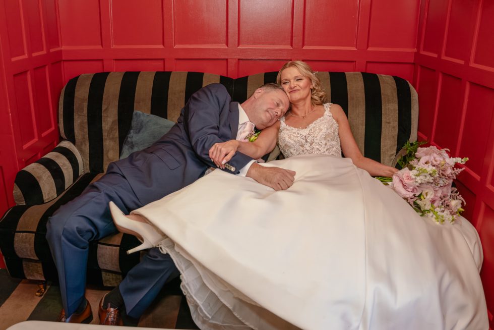 A relaxed bride and groom reclining on a striped sofa against red wood panelled walls at The Cromwell Hotel in Stevenage. The groom is resting his head on the bride's shoulder while she smiles contentedly, holding her pink bouquet.