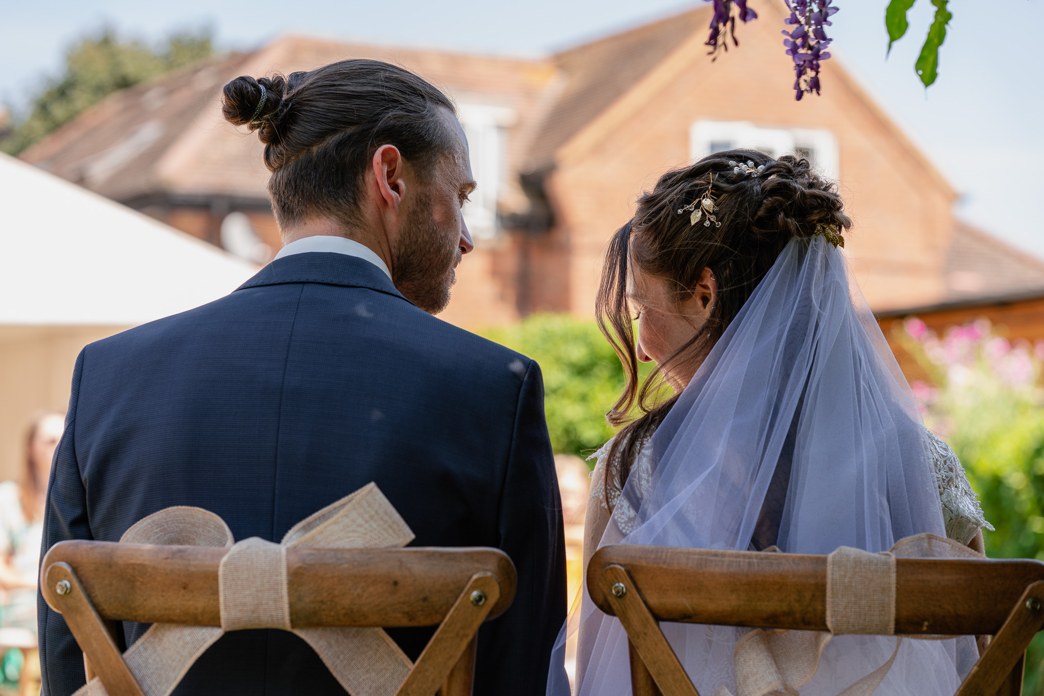 Bride and groom seated from behind sharing a glance during outdoor garden wedding ceremony, veil detail.