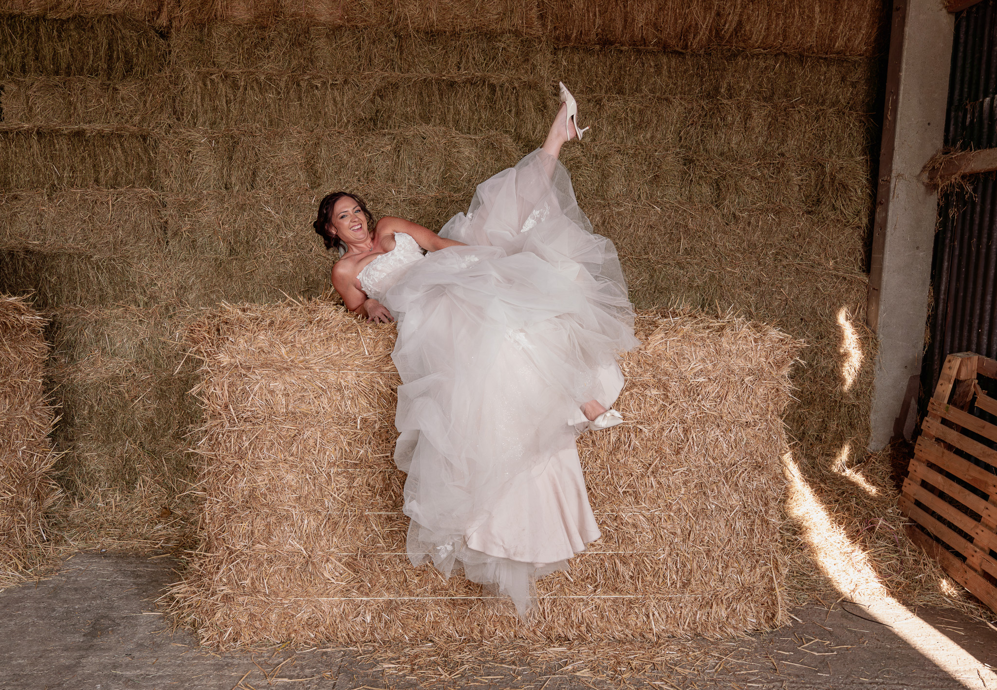 Fun Bride Portrait on Hay Bales | Barn Wedding Photography Hertfordshire Bride laughing and playfully kicking up her leg on hay bales in barn, fun candid wedding photography Hertfordshire