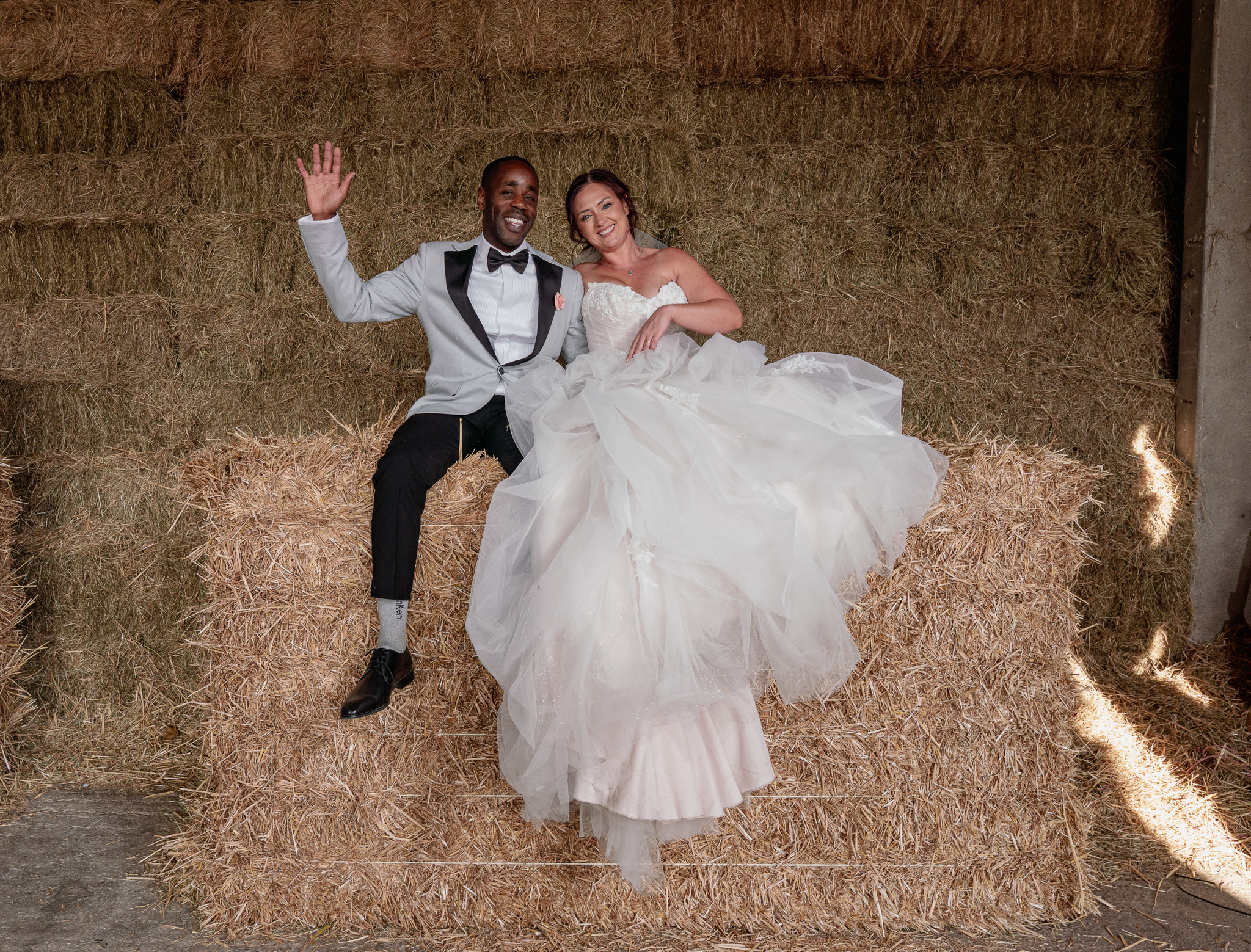 Fun Newlywed Portrait on Hay Bales | Barn Wedding Photography Hertfordshire Bride and groom laughing and waving on hay bales during fun, relaxed wedding portraits in Hertfordshire barn