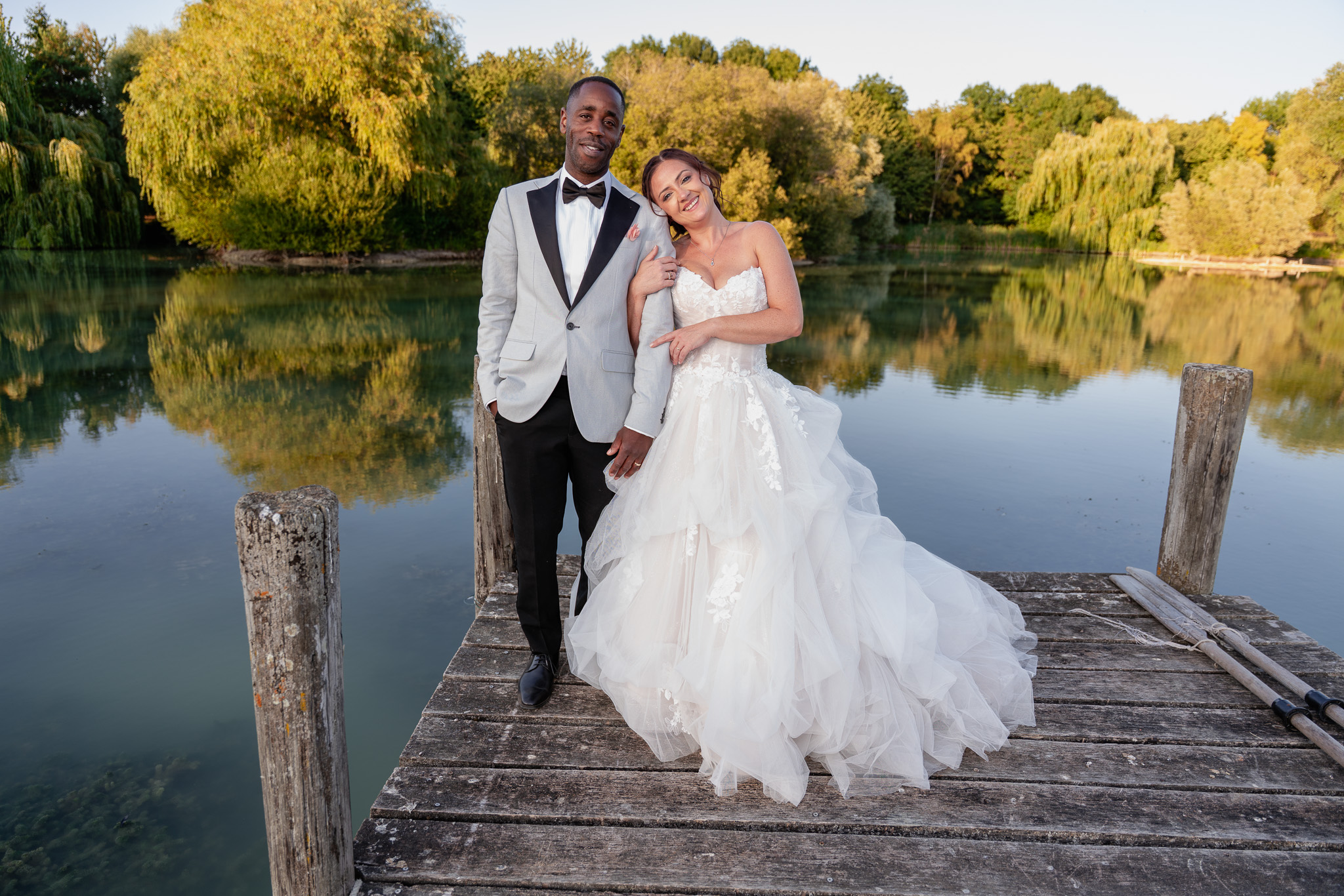 Bride and groom standing on wooden jetty by lake at golden hour, romantic wedding portrait Hertfordshire