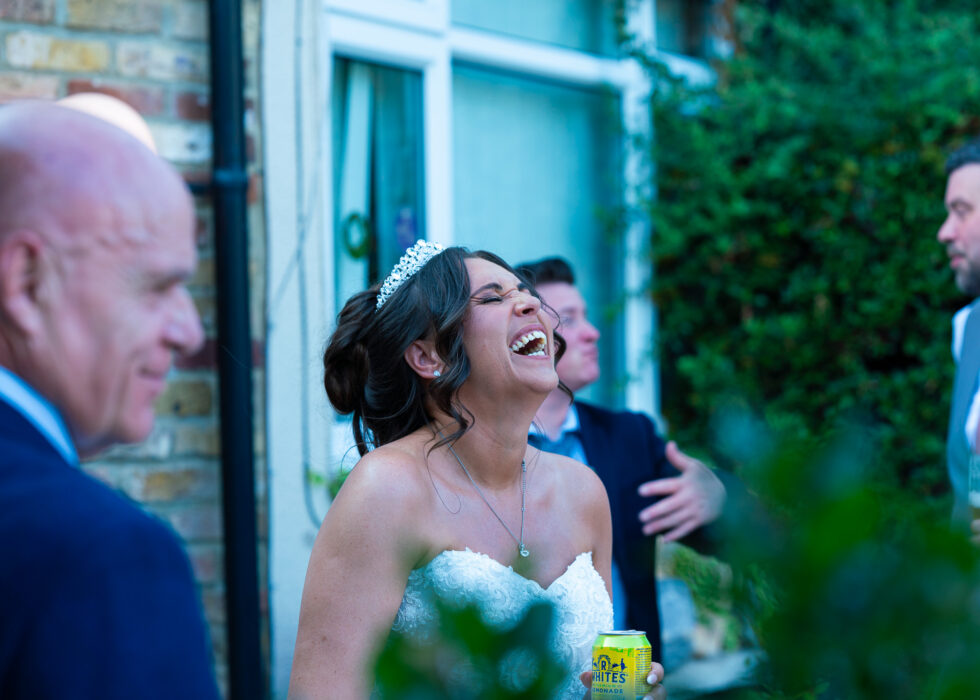 Bride with her head thrown back laughing hard outside a brick house while a guest in a navy suit stands in the foreground