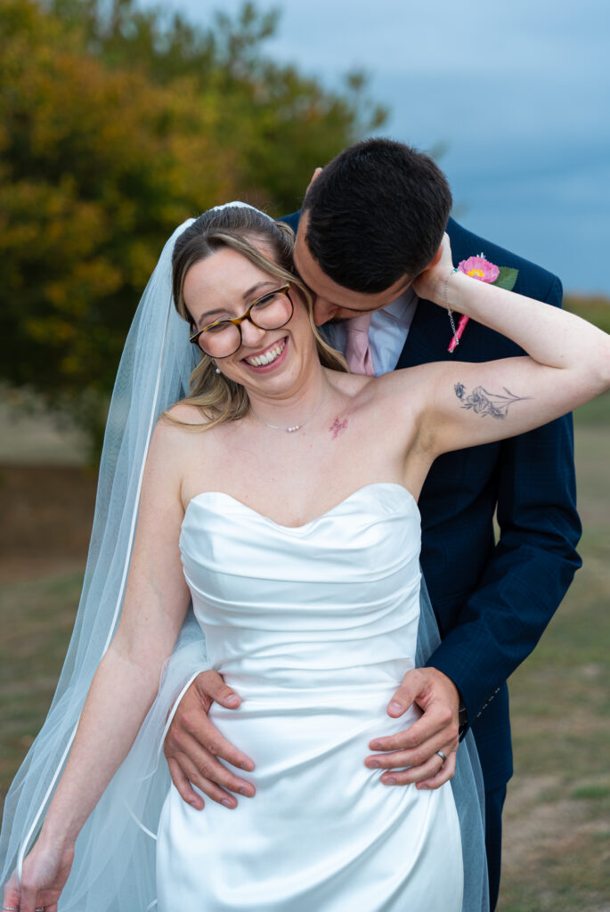 Bride in glasses and a long veil laughing with her head tipped back as her groom kisses her neck, his hands on her waist, against a soft autumn background