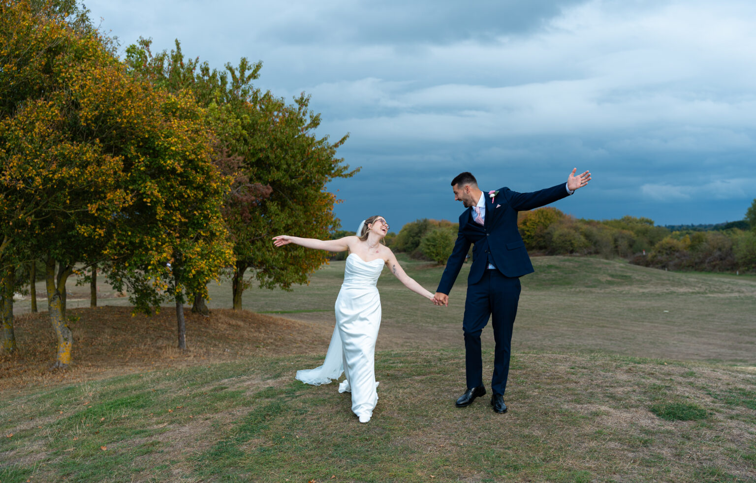 Bride and groom holding hands in a field, flinging their arms out and dancing under a stormy autumn sky