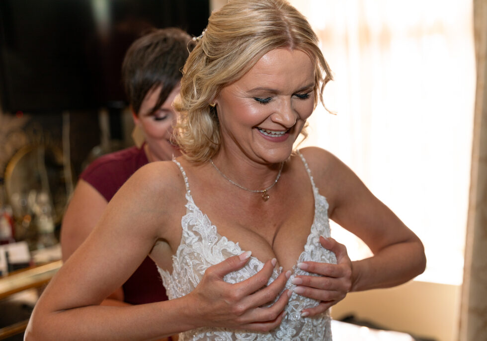 Bride in a lace dress laughing and looking down while a bridesmaid fastens the back
