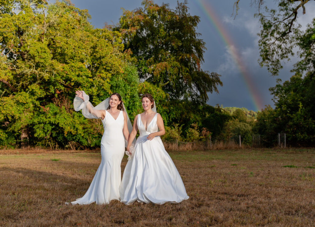 Two brides in white dresses walking hand in hand across a field, one waving her sheer sleeve, with a rainbow over the trees behind them