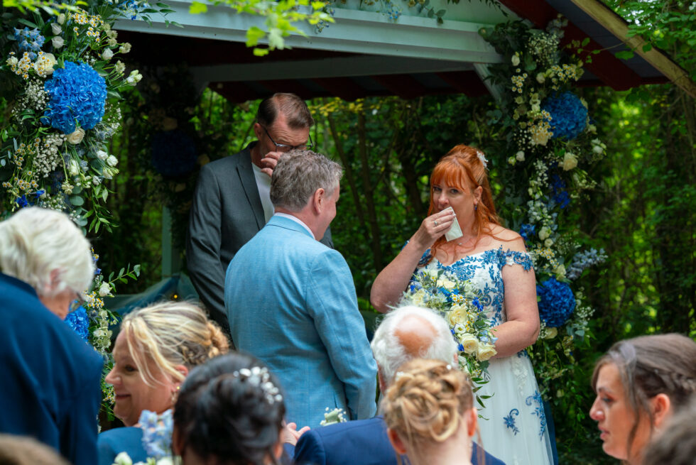 Bride and vicar both wiping away tears during the blessing at an outdoor wedding ceremony under a blue floral arch at the Sadie Centre, Enchanted Gardens