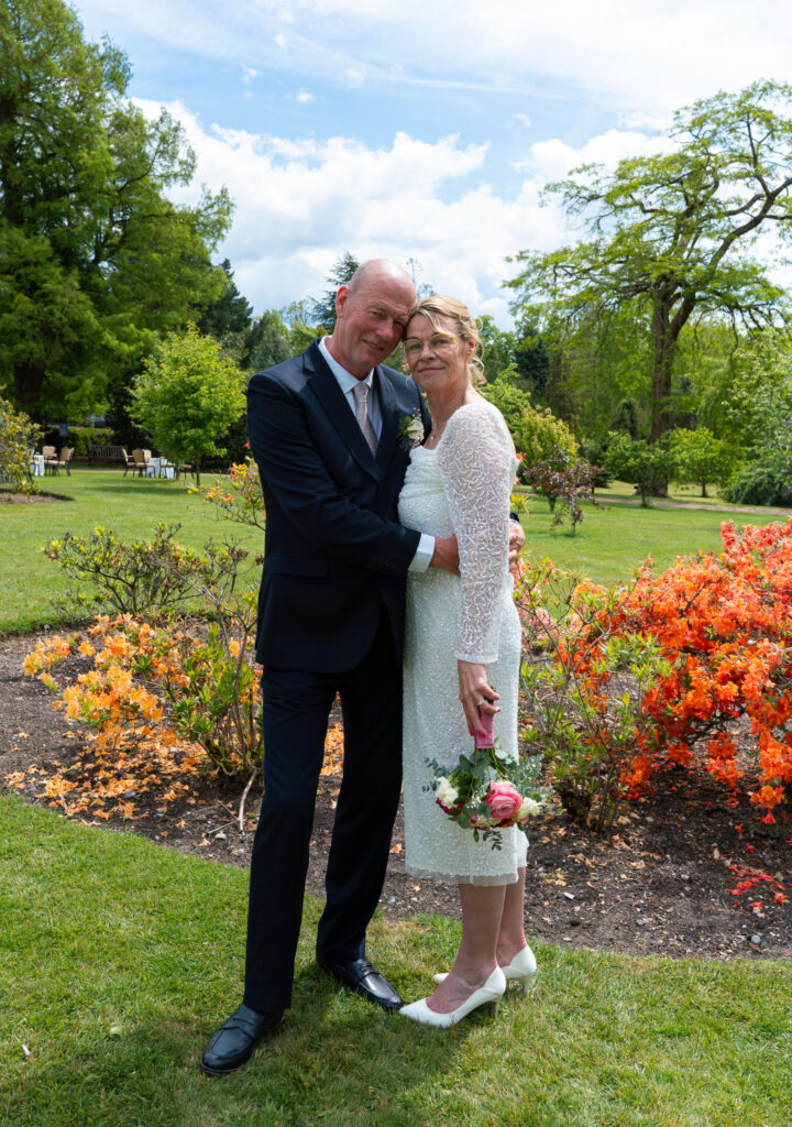 Older couple embracing in a garden on their wedding day, he in a navy suit and she in a white lace dress holding a pink bouquet, with orange azaleas beside them