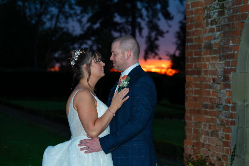 Bride and groom close together, face to face, against a glowing orange sunset beside an old brick wall at Fanham Hall, Ware