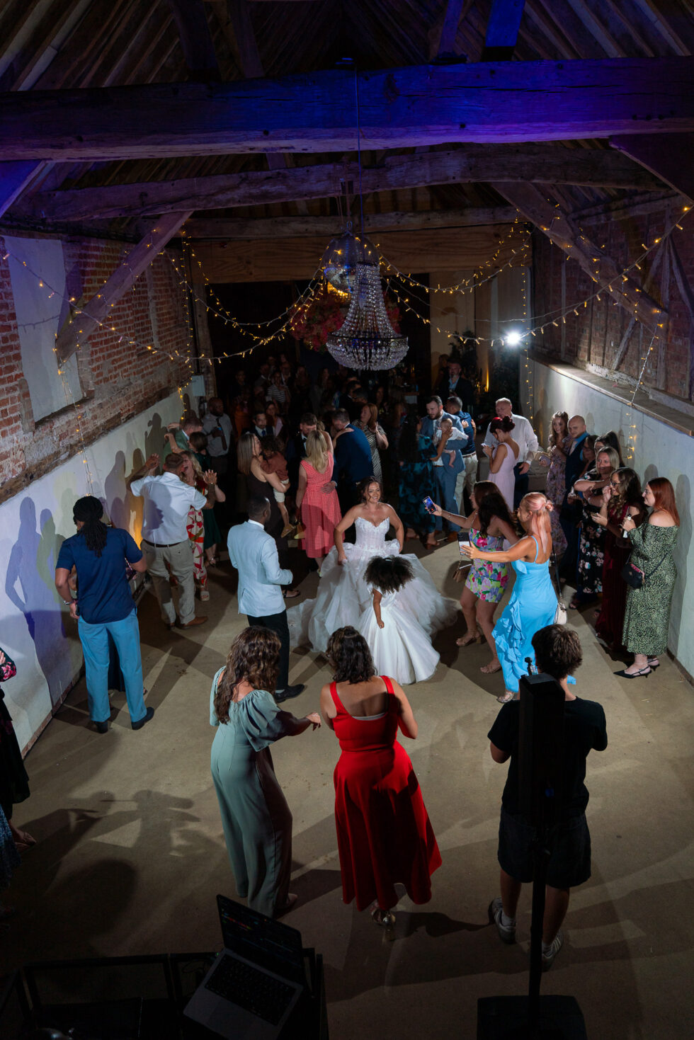 Bride in her wedding dress dancing with guests under fairy lights and a chandelier at the evening reception in the barn at Brook End Green Farm