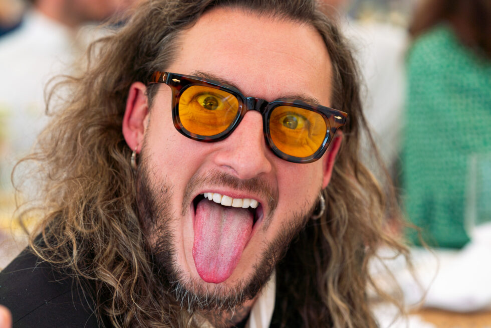 Wedding guest with long hair and orange tinted sunglasses sticking his tongue out at the camera