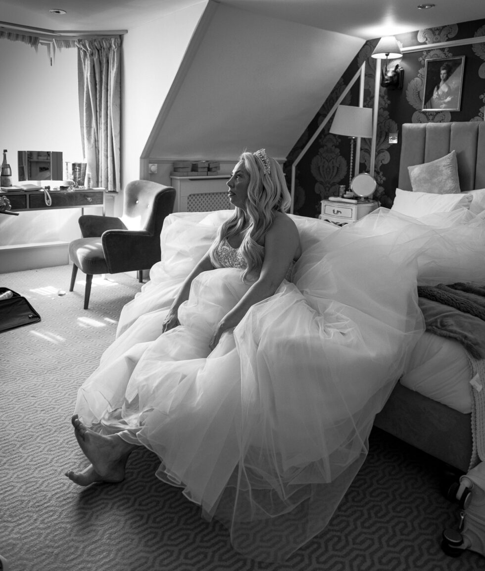 Black and white photo of a bride in her wedding dress and tiara sitting on the edge of the bed in her room at the Cromwell Hotel, Stevenage, a quiet moment before the ceremony