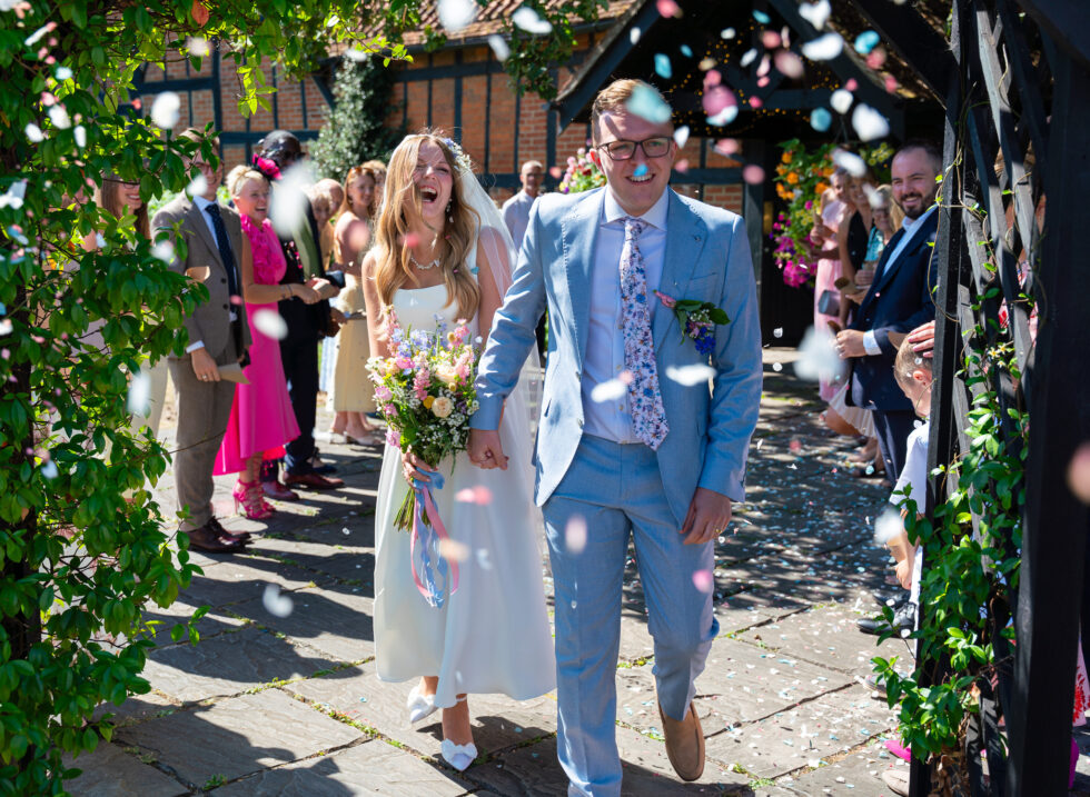 Bride and groom walking hand in hand through a confetti shower, laughing, with guests lining the courtyard at The Kingfisher, Bedford