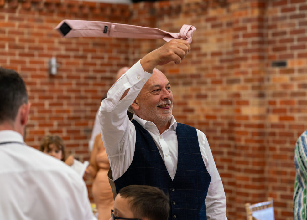 Father of the bride in a waistcoat swinging his tie above his head, cheering the newly married couple into their wedding breakfast at Eversholt Hall