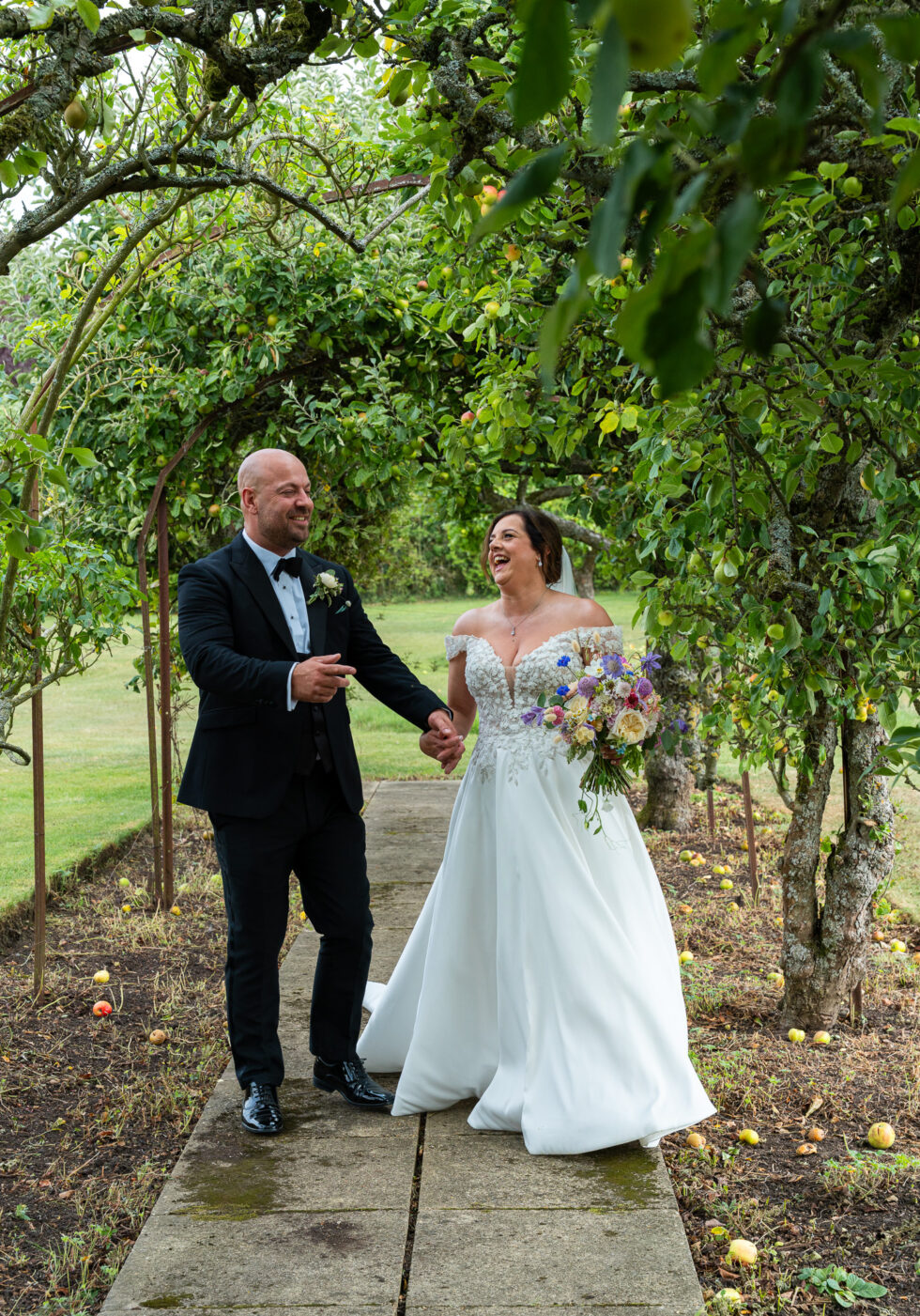 Bride and groom walking hand in hand and laughing under the fruit trees in the grounds of The Barns at Alswick, Hertfordshire
