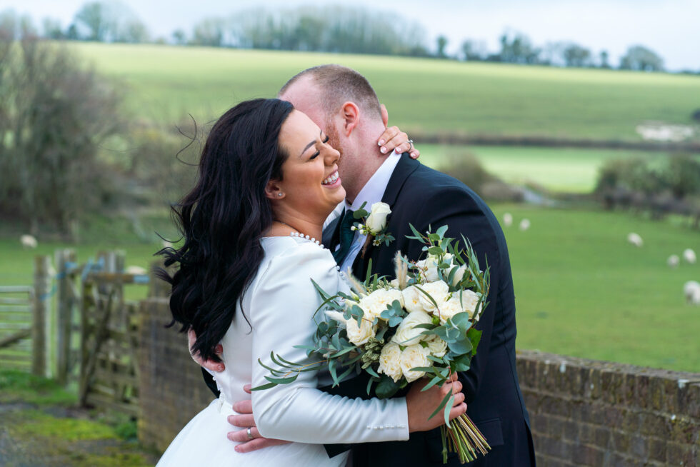 Bride in a long-sleeved dress and groom kissing in a green field holding a bouquet