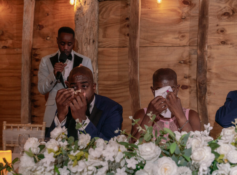 The groom's brother and mum wiping away tears at the top table while the groom gives his speech in the barn at Brook End Green Farm