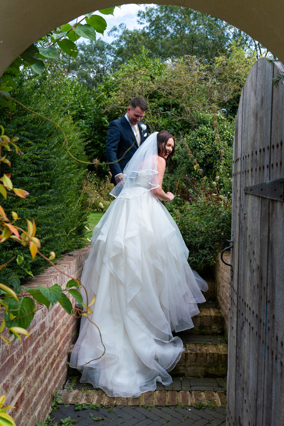 Bride in a full ruffled wedding dress walking down brick garden steps while the groom looks back at her