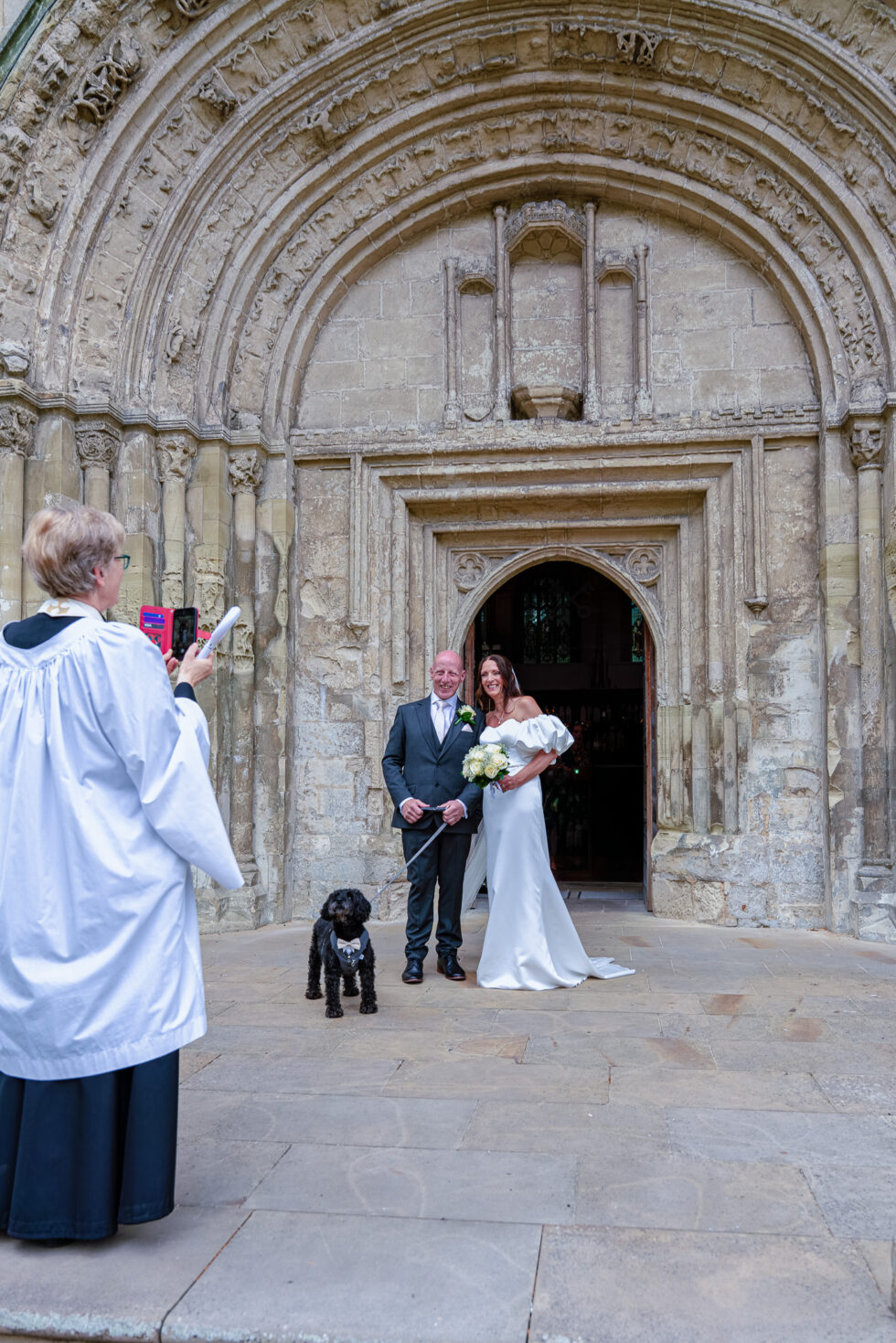 Bride and groom standing in an ornate stone church doorway with a small black dog while a robed vicar takes a photo