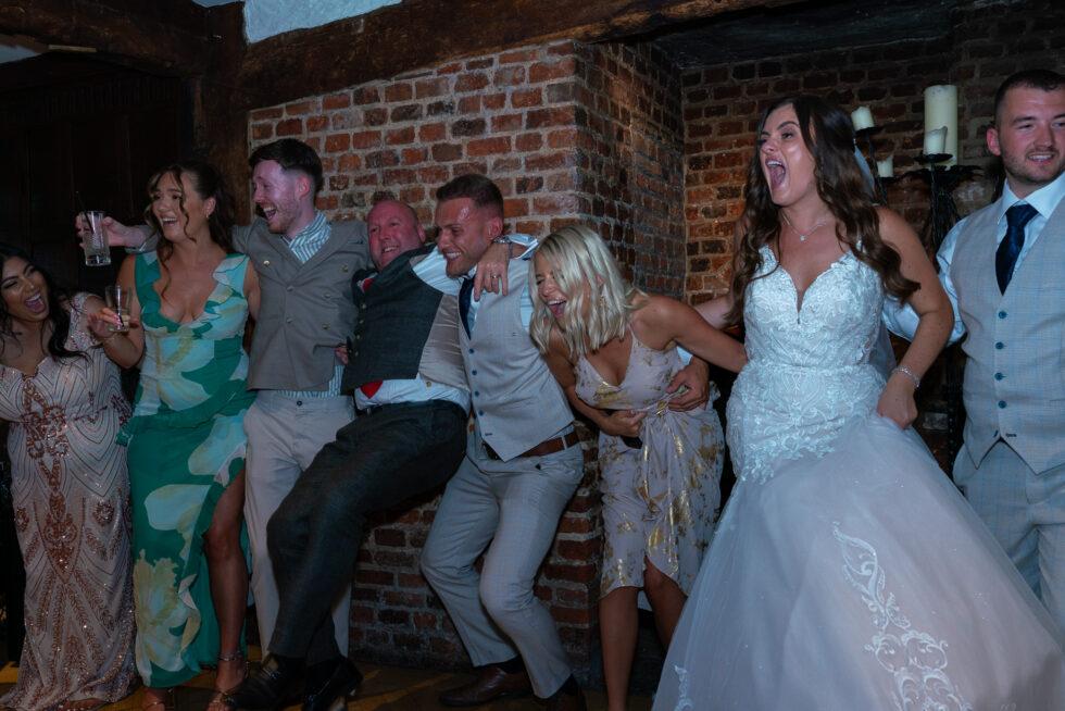Bride and groom dancing with their wedding guests, arms linked and laughing, in the brick-walled evening reception at Great Fosters Hotel