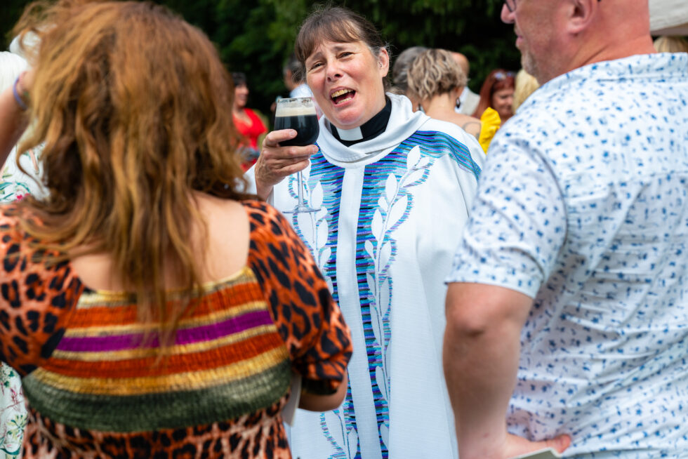 Vicar in white and blue robes raising a pint of dark beer and chatting with guests, one in a leopard print top in the foreground