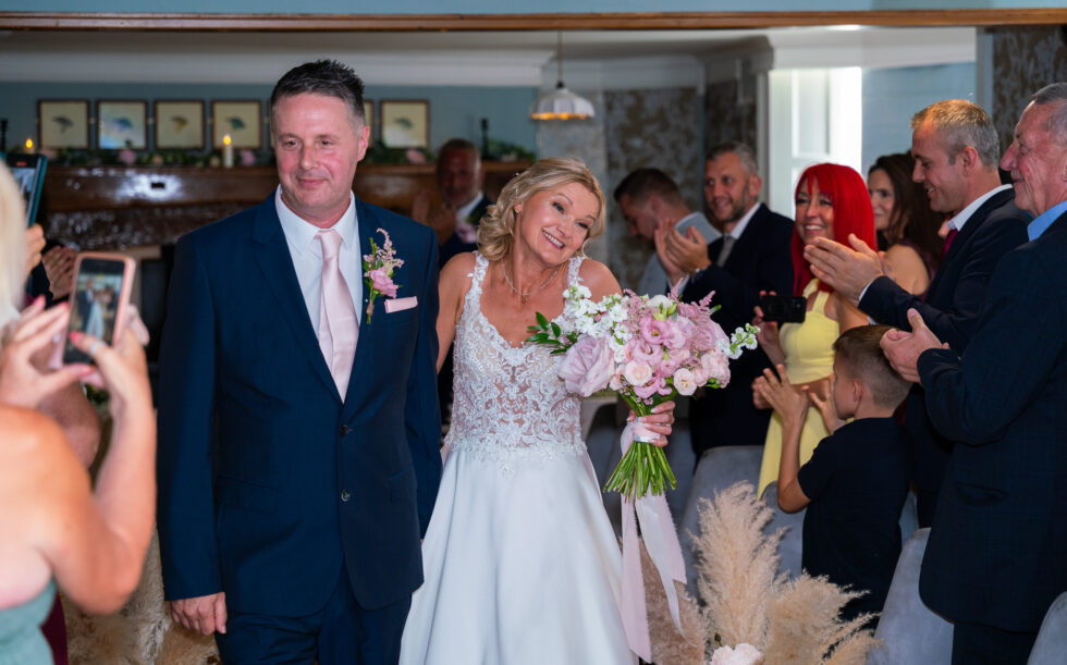 Bride and groom walking back down the aisle past clapping, phone-wielding guests after their ceremony at the Cromwell Hotel, Stevenage