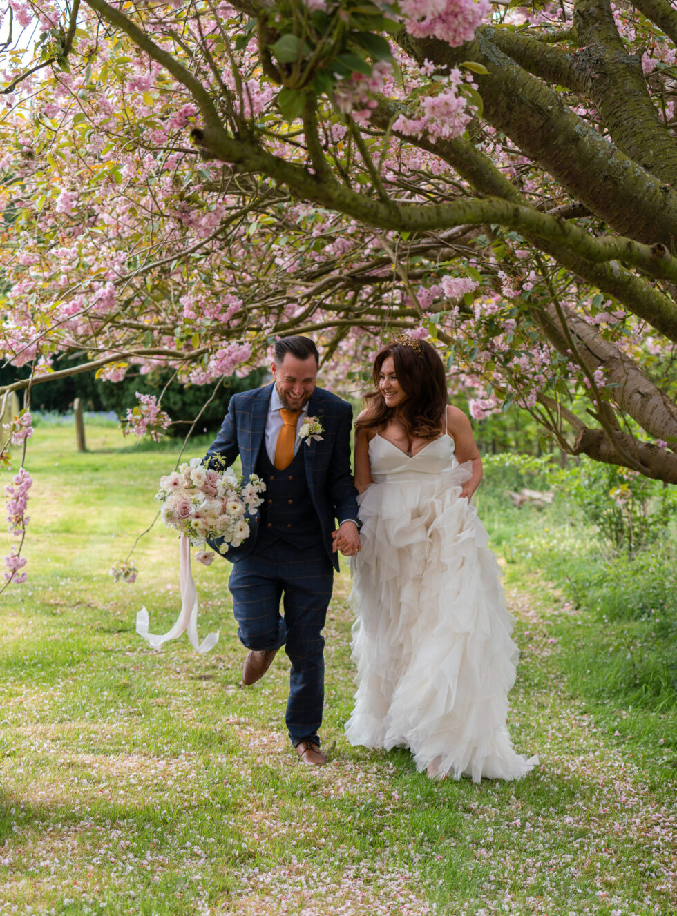 Groom in a navy suit and bride in a long dress walking through grass under pink cherry blossom
