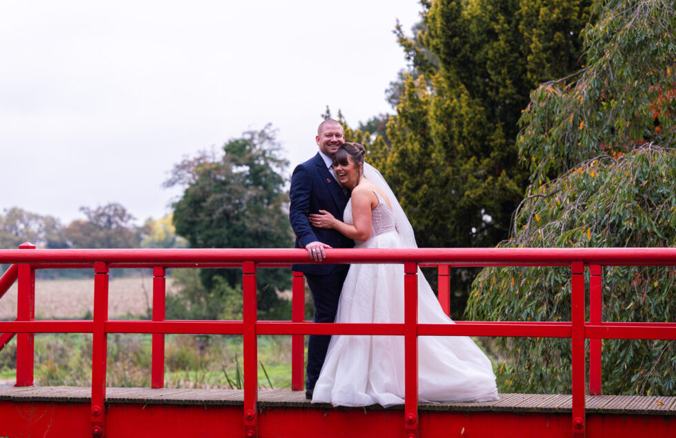 Bride and groom hugging on a bright red bridge in the gardens of Fanham Hall, Ware